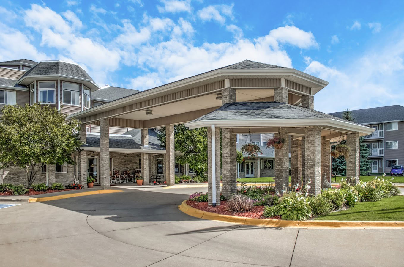 Entrance of The Mallard Point Senior Living with covered driveway, brick columns, landscaped garden, and blue sky.