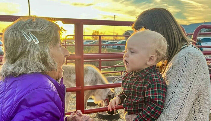 A senior resident in a purple jacket interacts joyfully with a baby held by a woman near a petting zoo, as the sun sets in the background, creating a warm and heartwarming moment.