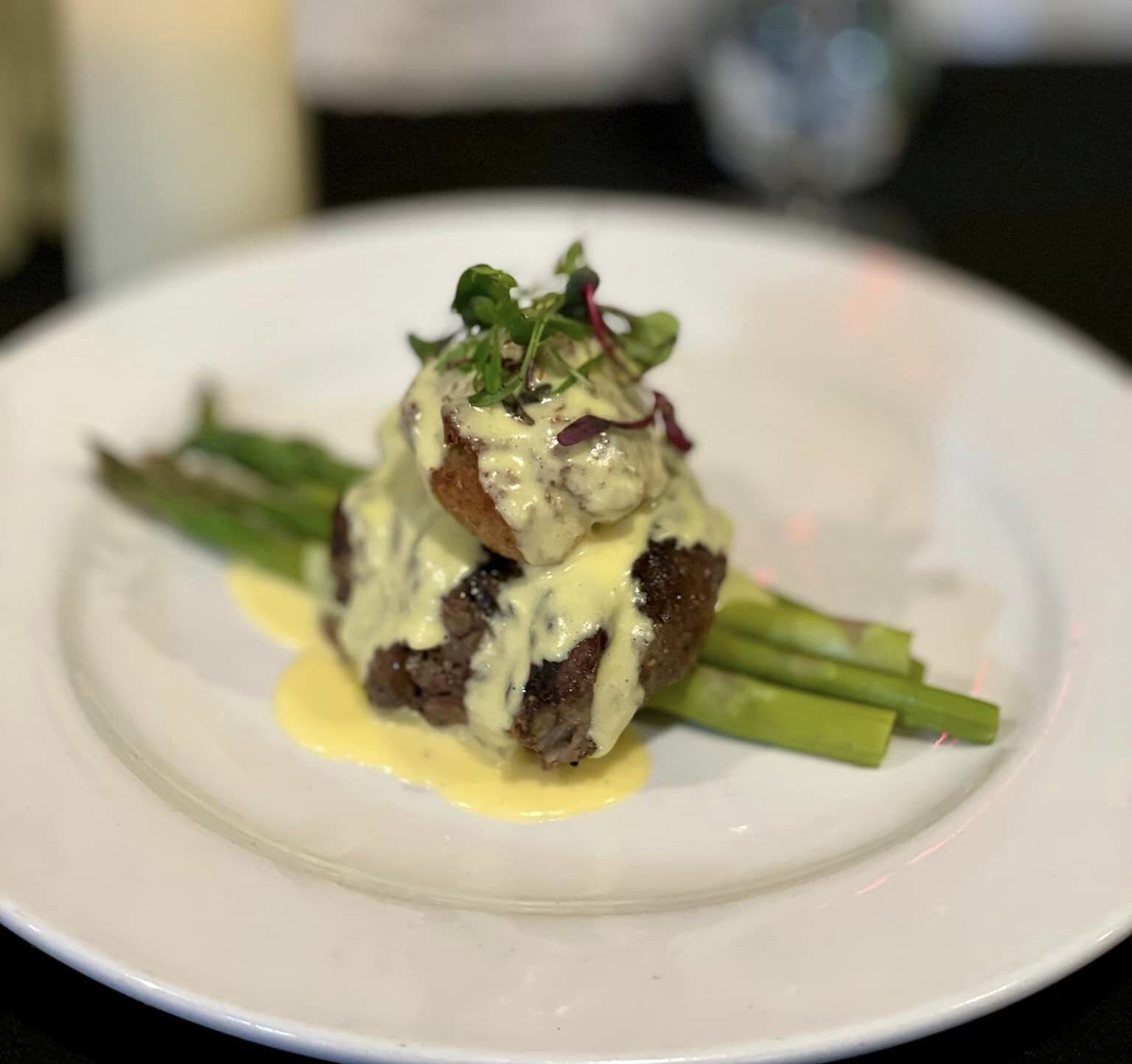 Close-up of a gourmet dish featuring crispy pork belly garnished with microgreens and accompanied by sautéed vegetables, presented on a plate with artistic sauce drizzles.