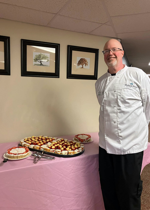 Chef in a white Mallard Point jacket standing next to a pink-covered dessert table filled with mini cheesecakes, fruit tarts, and assorted pastries.