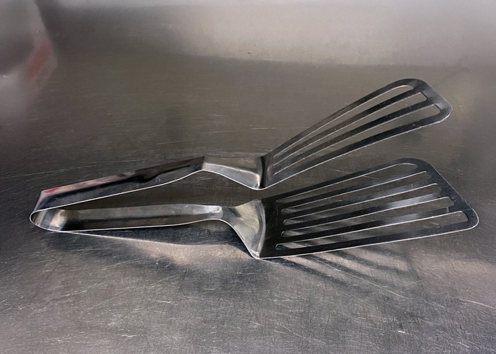 Metal double-spatula grabber tool resting on a stainless steel countertop, ideal for flipping thick sandwiches, burgers, and grilled items with precision.