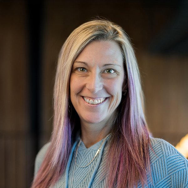 Nikki Morrissey, Senior Living Director at Mallard Point Senior Living, smiling in a light blue sweater with long blonde and purple hair against a softly lit background.