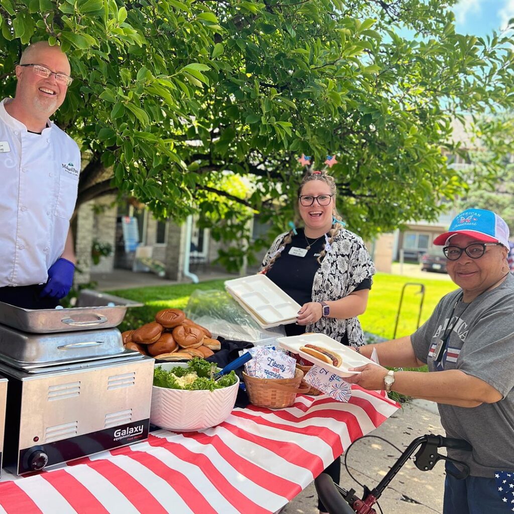 Culinary team member and staff serve fresh hot dogs and buns at an outdoor cookout with a resident, enjoying food and fellowship under a sunny tree.