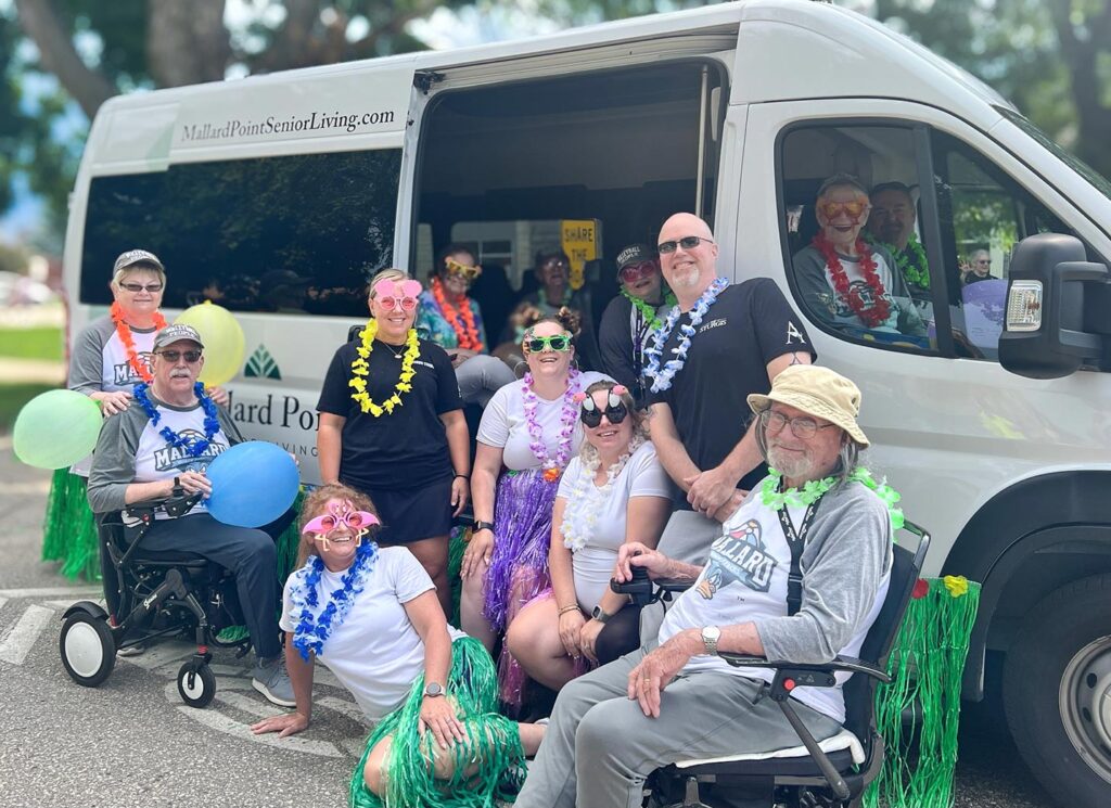Residents and team members from Mallard Point Senior Living dressed in leis and fun glasses pose by a community bus, ready to celebrate together outdoors.