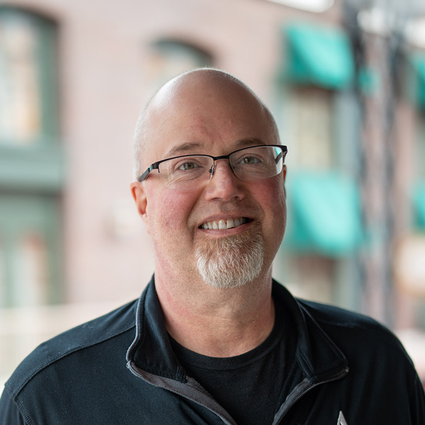 Kale Crawford, Culinary Director at Mallard Point Senior Living, smiling in a professional headshot, wearing glasses and a black zip-up jacket, with a softly blurred outdoor background.