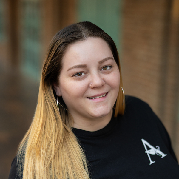 Ayron Appleby, Executive Director at Mallard Point Senior Living, smiling in a professional outdoor headshot with long ombré hair, hoop earrings, and a softly blurred brick background.