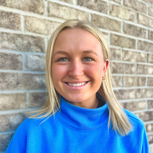 Cora Syverson, Senior Living Coordinator at Mallard Point Senior Living, smiling in a professional headshot, wearing a bright blue sweater, with a brick wall background.