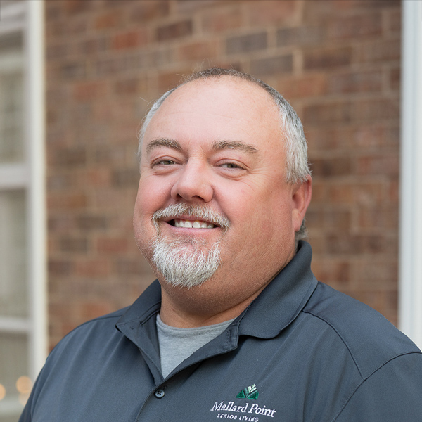 Bill Sager, Plant Operations Director at Mallard Point Senior Living, smiling in a professional outdoor headshot with short gray hair, a gray goatee, a dark polo shirt, and a softly blurred brick background.